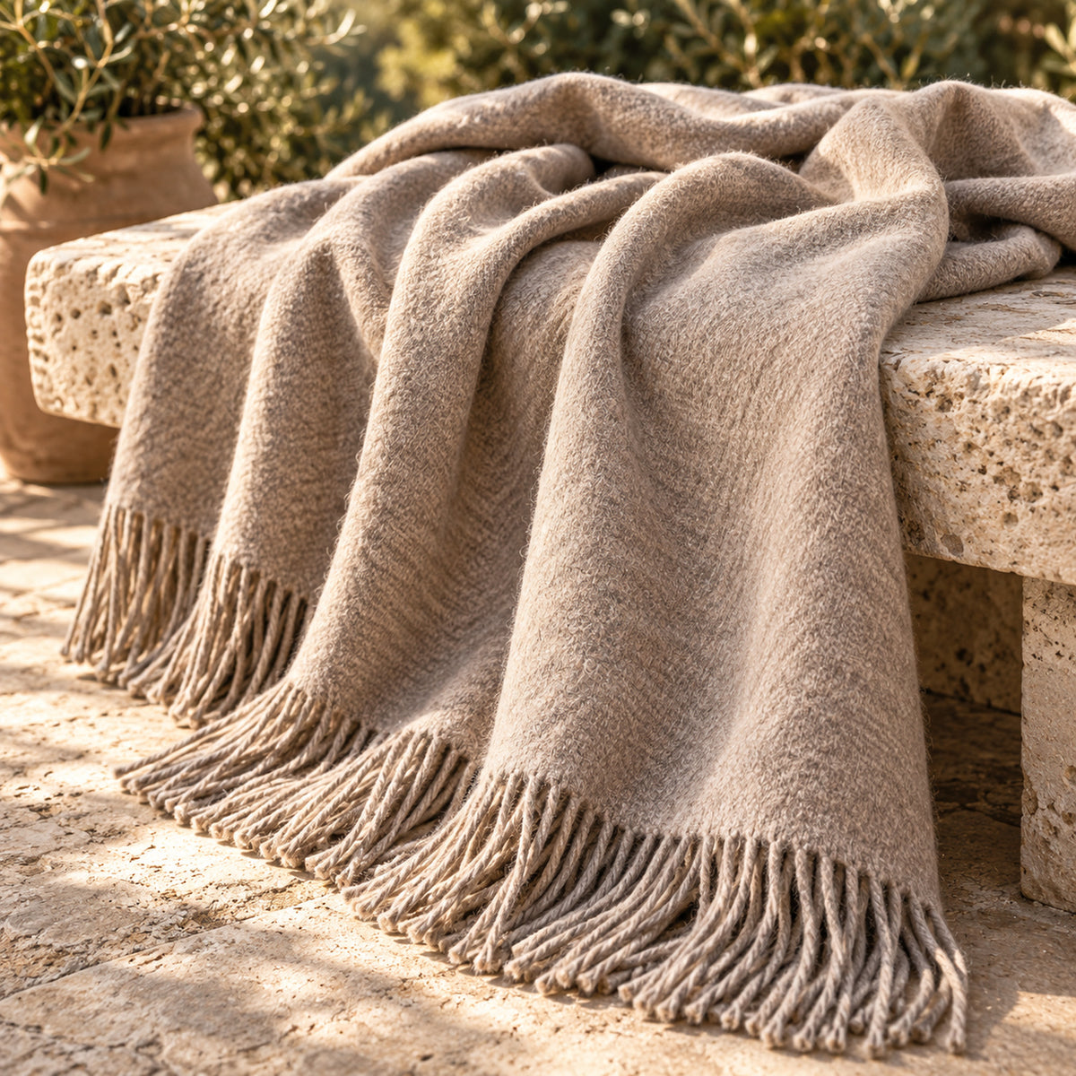 Un plaid en laine beige à franges, posé sur un banc en pierre à l'extérieur, la lumière du soleil mettant en valeur sa texture, avec des plantes en pot en arrière-plan.
