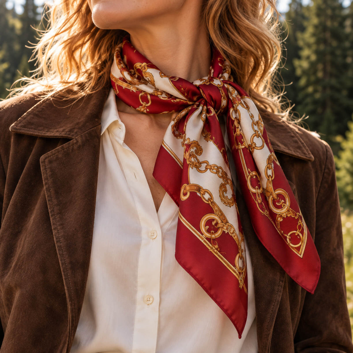 Femme vêtue d'une veste marron, d'un chemisier blanc et d'un foulard en soie rouge orné de motifs de chaînes dorées, debout à l'extérieur avec des arbres en arrière-plan.